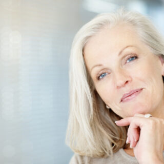 A woman with grey hair stares forward with a slight smile and her head tilted, resting on her hand which has a large ring on it.