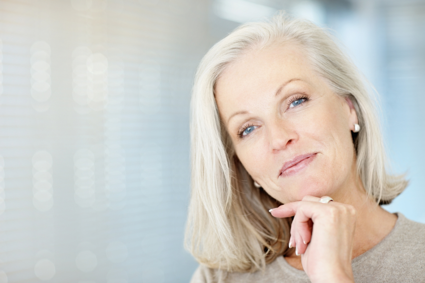 A woman with grey hair stares forward with a slight smile and her head tilted, resting on her hand which has a large ring on it.